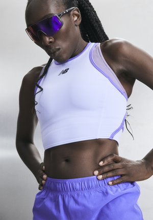 Atletická osoba s vrkočmi, ktorá nosí fialové reflexné športové slnečné okuliare, biely a svetlofialový bezrukávový crop top a fialové krátke nohavice.