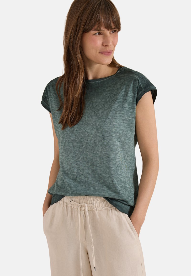 Femme aux longs cheveux bruns portant un haut vert foncé à manches courtes et un pantalon beige à cordon, debout les mains dans les poches, regardant vers la droite.