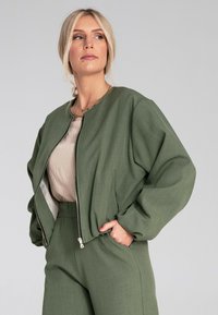 Zielona bomberka o gładkiej fakturze, z ribbowanym dekoltem i zapięciem na zamek, w połączeniu z jasnobeżową bluzką i dopasowanymi spodniami z wysokim stanem.