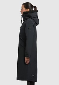 Parka neagră lungă cu glugă, având un guler căptușit cu fleece, buzunare cu fermoar și un patch cu logo pe lateral. Material neted și gros.