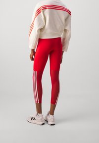 Červené legíny s síťovinovými vkládacími díly a bílými pruhy, zkombinovan é se krémovou mikinou s červenými pruhy po ramenou, nošeny s bílými teniskami.