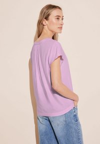 Lila kortärmad topp med en avslappnad passform, rund halsringning och subtil detalj med veck bak, kombinerad med ljusblå denim jeans.