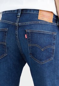 Vue de dos d'une personne portant un jean Levi's 501 bleu avec coutures visibles sur les poches, étiquette rouge et patch en cuir marron de la marque sur la ceinture.