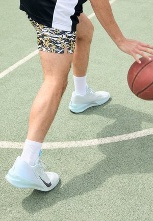 Buty do koszykówki w jasnym miętowym kolorze z teksturowanym materiałem, zestawione z czarno-żółtymi szortami w wzory, na boisku sportowym.