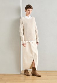 Beżowy sweter w prążki, biały koszulowy podkoszulek, kremowa spódnica midi z rozcięciem z przodu, brązowe zamszowe botki za kostkę, na jasnym tle.