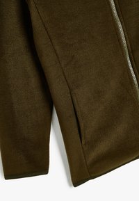 Kurtka polarowa w kolorze oliwkowym z widocznym rękawem i szwem bocznym, srebrnym zamkiem błyskawicznym oraz bocznym otworem kieszeni, na białym tle.