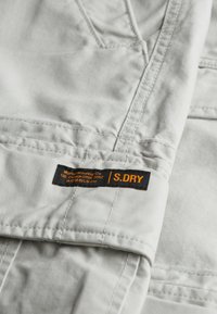Lys grå stof med synlige sømme; har et sort etiket med orange tekst, der står "S.DRY" og yderligere produktionsdetaljer.