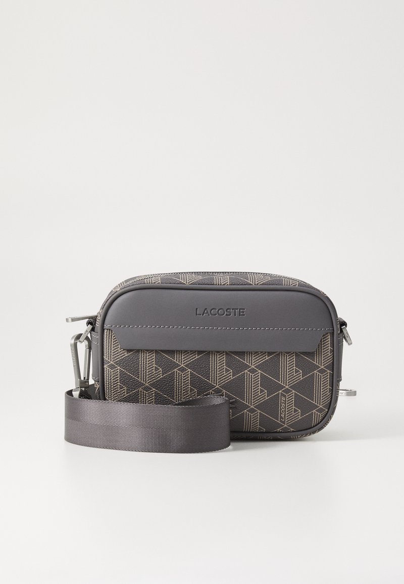 Grå mønstrete crossover-veske med en strukturert overflate, med en glatt grå stropp og en sølvglinsende glidelås. Merkevaren er preget på forsiden.