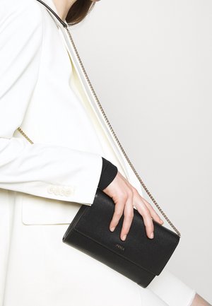 Clutch in pelle nera con finitura testurizzata e tracolla a catena dorata, tenuta sotto un blazer bianco. Design semplice con una silhouette pulita.