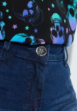 Top nero con motivo colorato di teschi e stelle, abbinato a jeans a vita alta blu scuro con dettaglio di bottone a forma di stella argentata.