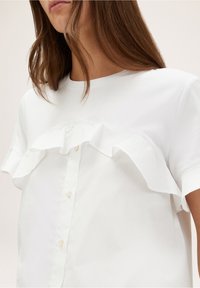 Blouse blanche avec un volant au niveau de la poitrine, des manches courtes et une fermeture à boutons sur le devant. Tissu doux avec un design épuré et minimal.