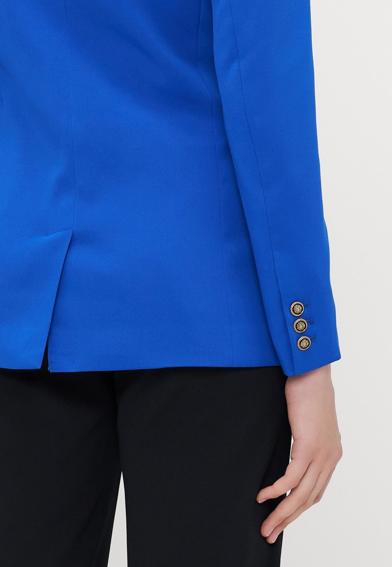 Blazer ajusté bleu avec une texture lisse et trois accents de boutons en or au niveau du poignet. Présente une légère fente à l'arrière.
