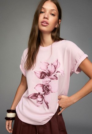 Light pink short-sleeve t-shirt with a floral design in burgundy, featuring rolled sleeves and a loose fit, paired with brown pants and accessories.