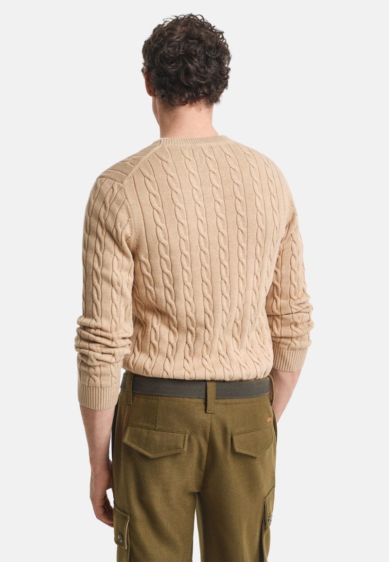 Beige Kabelstrickpullover mit strukturiertem Muster, gerippten Bündchen und rundem Halsausschnitt; kombiniert mit olivgrünen Hosen mit Taschen.