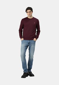 Maglione color bordeaux con scollo a girocollo e polsini a coste, abbinato a jeans azzurri e scarpe nere. Design semplice con un dettaglio ricamato.