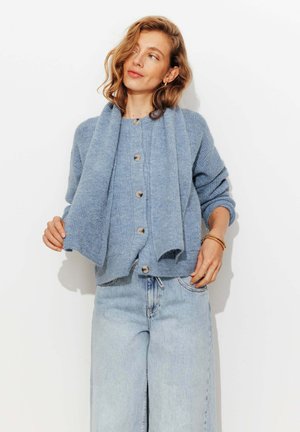 Une femme aux cheveux ondulés porte un cardigan bleu boutonné avec une écharpe assortie et un jean taille haute bleu clair, sur un fond blanc.