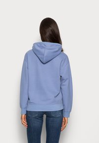 Kvinna med långt brunt hår som bär en ljusblå hoodie och mörkblå jeans, stående vänd mot en enfärgad vit vägg.