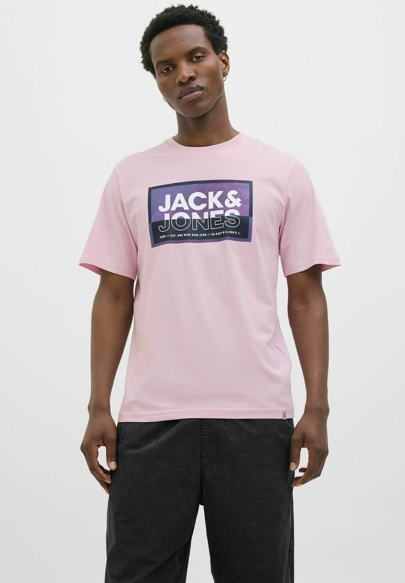 Lyserød bomulds T-shirt med et rektangulært grafisk design med "JACK & JONES" tekst. Kortærmet og rund hals, parret med mørke bukser.
