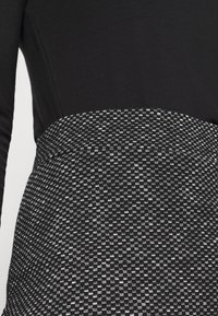 Schwarzes Langarmoberteil, das mit einem schwarz-weiß strukturierten Rock verbunden ist, der ein Muster aus abwechselnden Quadraten aufweist und eine glatte Stofftextur betont.