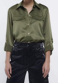 Olivgrön satinblus med långa ärmar, två framfickor och gyllene knappar. Buren med mörka, strukturerade byxor för kontrast.