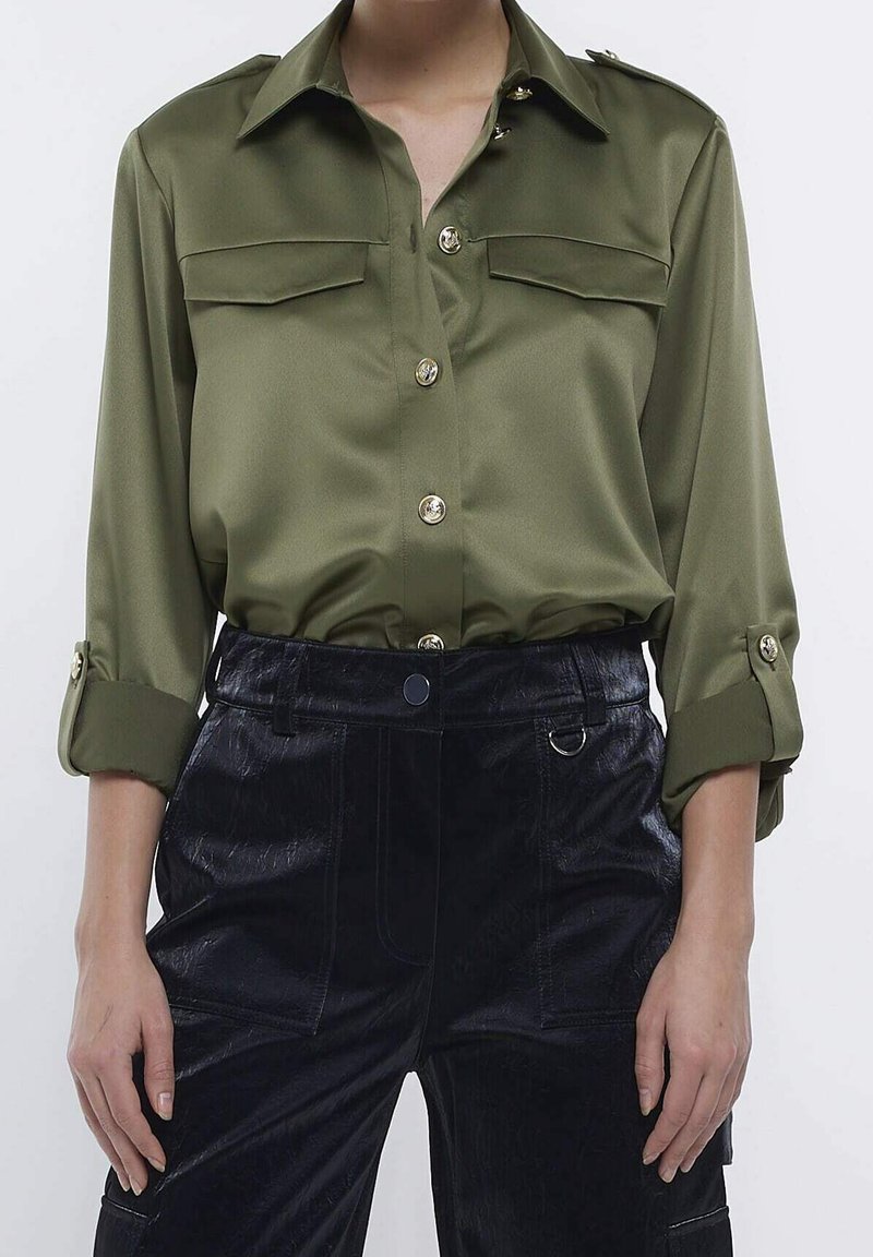 Olivgrön satinblus med långa ärmar, två framfickor och gyllene knappar. Buren med mörka, strukturerade byxor för kontrast.