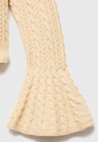Manica di un maglione lavorato a maglia color crema con motivo a trecce e fondo a campana su sfondo bianco.