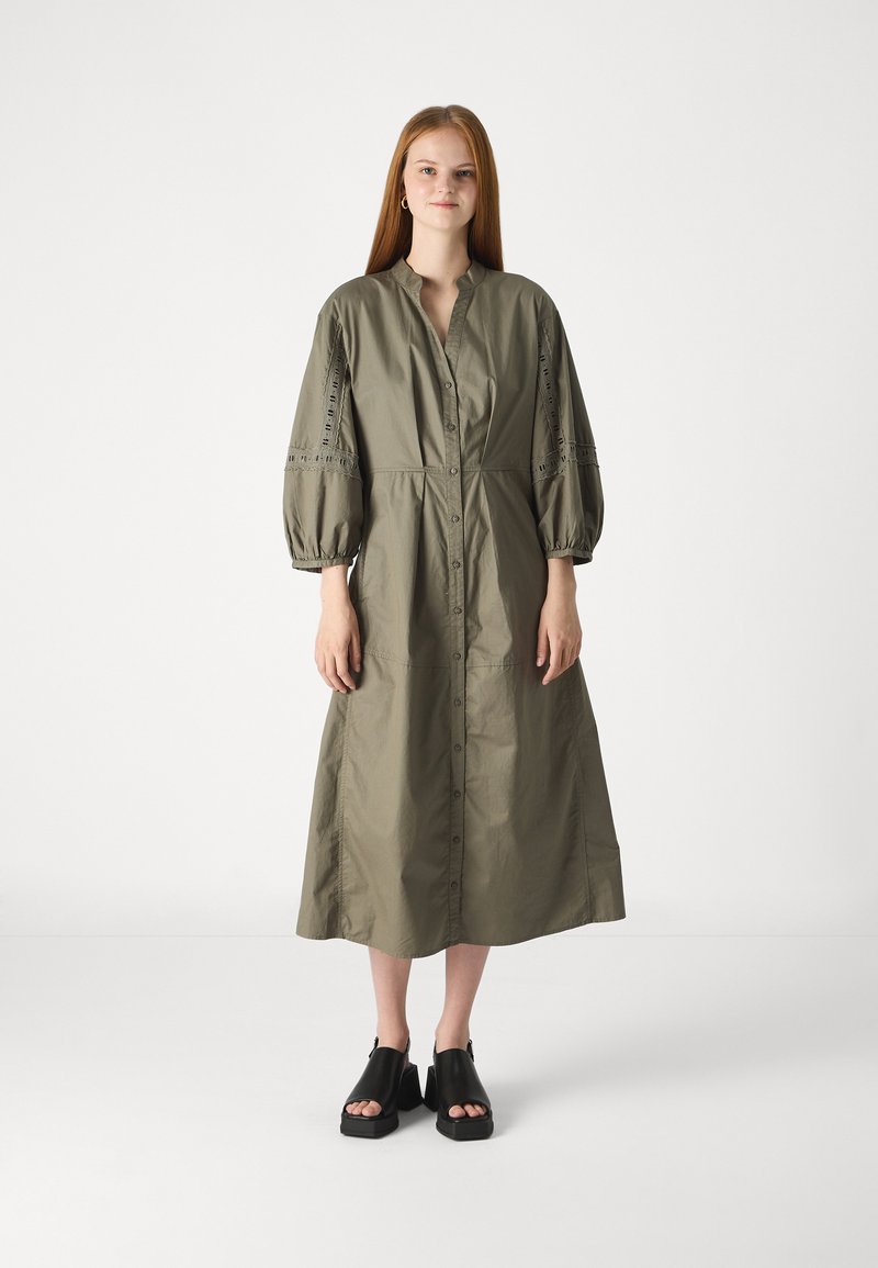 Vestito verde oliva, con bottoni, colletto, maniche a pallone e dettagli in pizzo. Abbinato a scarpe con zeppa nere. Tessuto morbido, lunghezza al ginocchio.