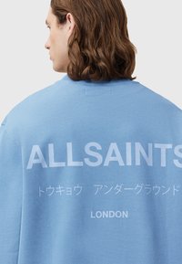 Modrá mikina vyrobená z bavlny, na zadní straně s bílým textem: „ALLSAINTS,“ japonské znaky a „LONDON.“ Volný, pohodlný střih.