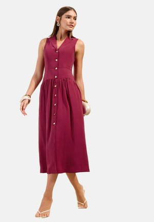 Kobieta ubrana w bezrękawową bordową sukienkę midi na guziki, białe sandały na obcasie i złoto-białe bransoletki, idąca na tle jednolitego tła.