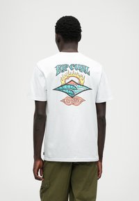 T-shirt de algodão branca com um design gráfico colorido na parte de trás, apresentando ondas, chamas e o texto "RIP CURL" acima.