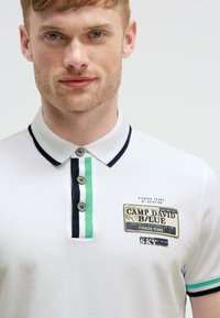 Weißes Poloshirt mit marineblauen und grünen Streifenakzenten, dreiknopfiger Knopfleiste und einem gestickten Logopatch auf der Brust.