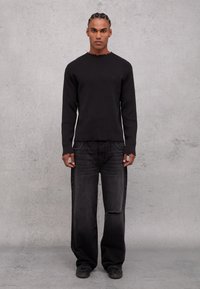 Maglietta nera a maniche lunghe con orlo grezzo, abbinata a jeans neri larghi con ginocchio strappato. Indossata con scarpe nere.