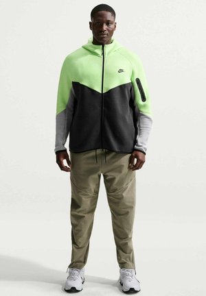 Uomo in piedi che indossa una felpa con cappuccio Nike verde neon, nera e grigia, pantaloni beige e sneaker bianche, su uno sfondo chiaro uniforme.