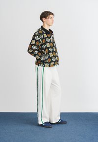 Svart hettejakke med blomstermotiv og logo; hvite vide bukser med grønn stripe; marineblå sko. Modellen poserer sidelengs mot en enkel bakgrunn.