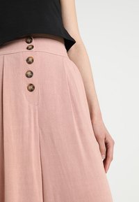 Pink skirt featuring a high waist, pleats, and five brown button accents down the front. Fabric texture appears smooth and lightweight.