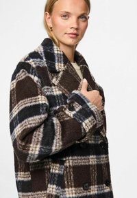 Dubbelzijdige geruite jas in bruin, crème en marineblauw; textiel met een gegolfde stof, met een gekliefde kraag en zwarte knopen. Model poseert met een opgestoken hand.