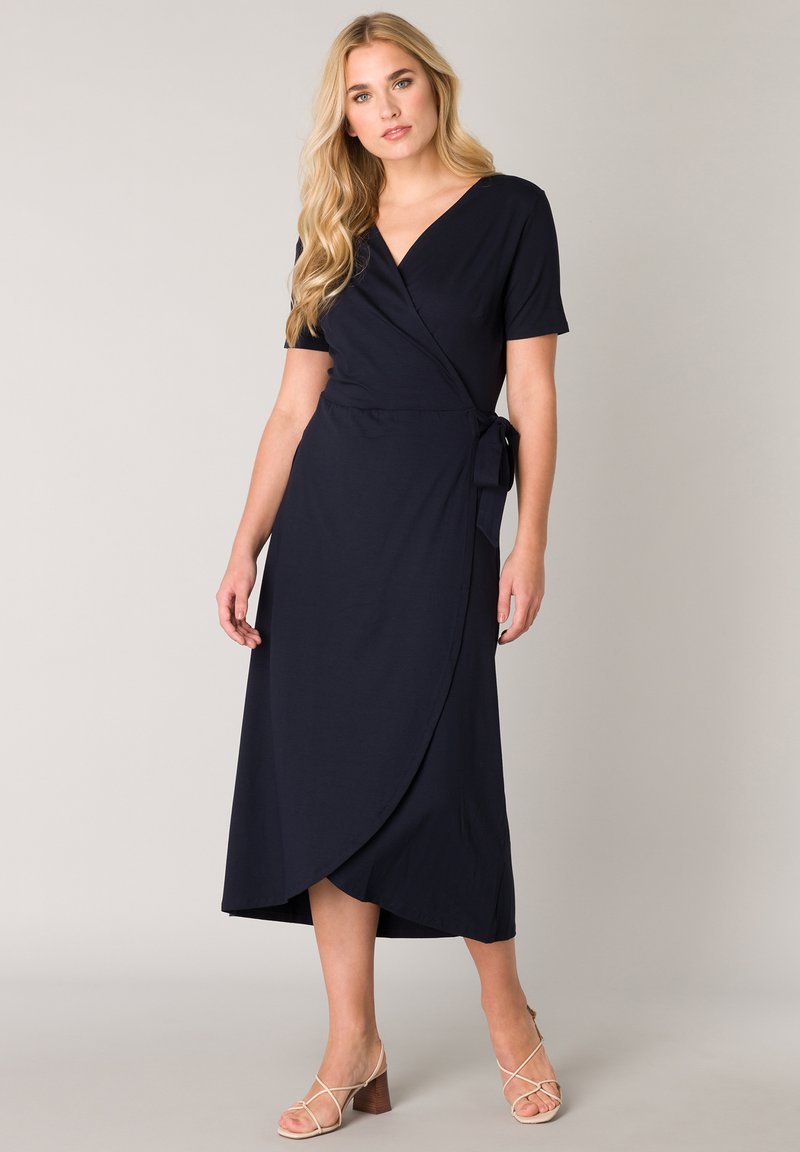 Marinefarbener Wickelkleid mit kurzen Ärmeln, glatter Textur, knielanger Saum, seitlicher Bindegürtel und ergänzt durch leichte Riemchen-Absatzsandalen.