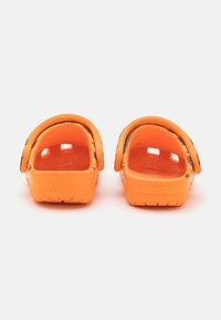 Orange træklipper med en struktureret overflade, åben bag og justerbar rem. Lavet af syntetisk materiale og har ventilationshuller for åndbarhed.