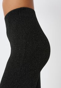 Leggings neri con una texture scintillante, vita alta e design aderente. Il materiale sembra elastico e liscio.