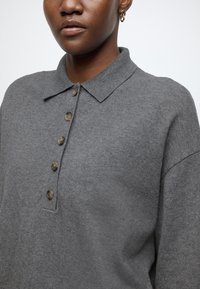 Tricou polo gri tricotat cu guler, prevăzut cu un guler cu șase nasturi și material texturat, afișat pe un fundal neutru.