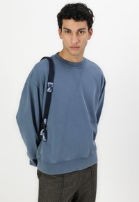 Sudadera azul con cuello redondo, puños acanalados y un pequeño logo bordado. Combinada con pantalones grises con patrón y una correa oscura.