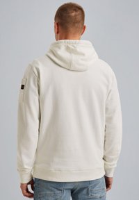 Svetlo béžová mikina s kapucňou na šnúrku, rebrovanými manžetami a spodným lemom. Má nášivku na ľavom rukáve a hladký, štruktúrovaný povrch.