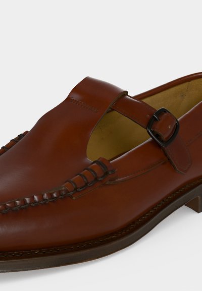 Chaussure habillée en cuir marron avec une sangle à boucle latérale, couture moc toe et un talon bas empilé sur fond blanc.