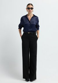 Chemise en satin bleu marine à boutons, portée avec un pantalon noir taille haute. Manches longues retroussées, ceinture noire avec accessoires dorés et talons noirs.