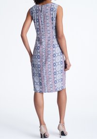 Robe sans manches avec un design ajusté, ornée d'un motif floral bleu et rose. Détail en forme de trou à l'arrière. Portée avec des chaussures à talons hauts couleur nude.