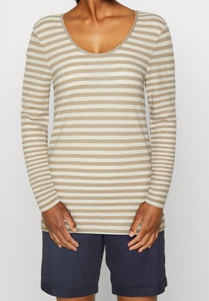 Osoba ubrana w beżowo-białą koszulę w paski z długim rękawem i dekoltem typu scoop oraz ciemnoniebieskie szorty, ręce spoczywają przy biodrach.