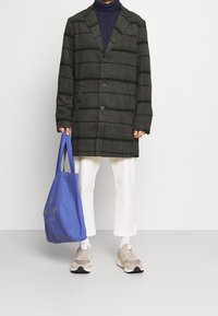 Grå ternet frakke med grønne striber, to knapper, sidelommer; marineblå turtleneck, hvide bukser, beige sneakers, holder en blå totebag.