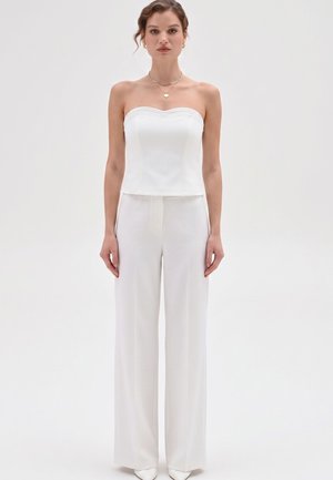 Femme portant un haut blanc sans bretelles et un pantalon blanc à jambes larges, debout devant un fond uni clair, face à l'avant.