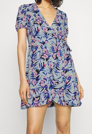 Woman wearing a short floral wrap dress with puff sleeves and blue, pink, and green leaf and flower patterns against a white background.