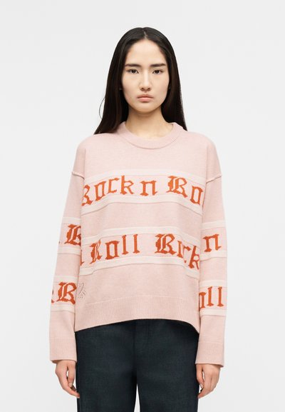 Jeune femme aux longs cheveux noirs portant un pull rose avec le texte orange « Rock n Roll » et un pantalon foncé, debout devant un fond blanc uni.